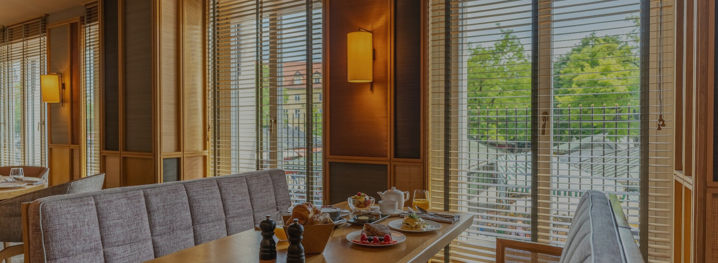 Gemütlicher Frühstücksbereich im LOUIS Hotel München mit gedecktem Tisch und Blick durch große Fenster mit Jalousien.