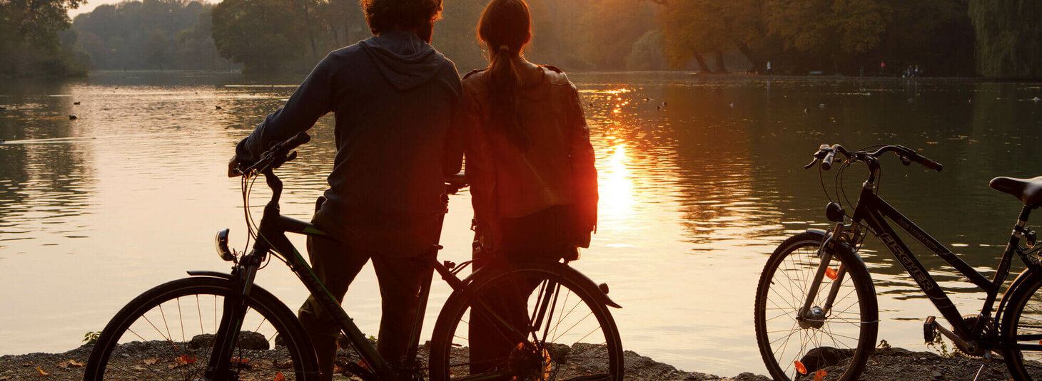 Zwei Menschen mit Fahrrädern stehen am Seeufer und genießen den Sonnenuntergang.