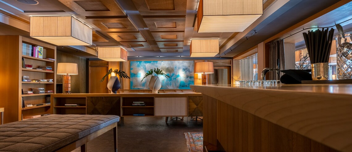 Moderne Hotel-Lounge mit Holzmöbeln, Bücherregal, stilvollen Lampen und Kunstwerk an der Wand im LOUIS Hotel München.