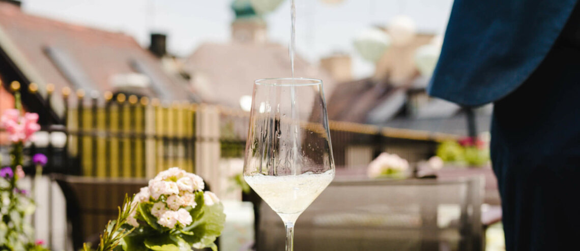 Weißwein wird auf einer sonnigen Hotelterrasse in ein Glas eingeschenkt, mit Blumen und Stadtblick im Hintergrund.