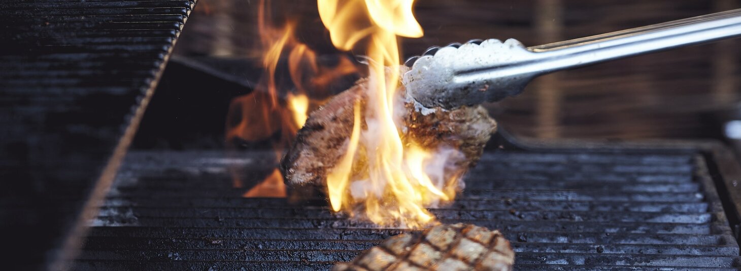 Steak wird auf einem Grill mit Flammen gegrillt, eine Zange hält das Fleisch.