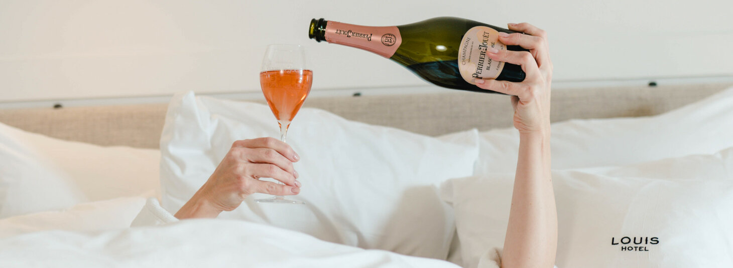 Person im Bett gießt roséfarbenen Champagner in ein Glas, LOUIS Hotel-Logo auf Kissen sichtbar.