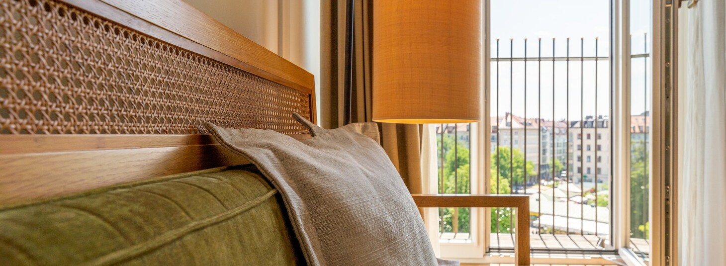 Gemütliches Hotelzimmer mit grünem Sofa, Kissen, hölzernem Kopfteil und Blick auf die Stadt durch offene Fenster.