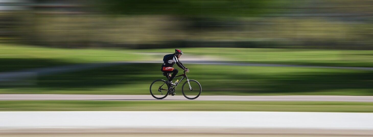 Radfahrer mit rotem Helm fährt schnell auf einem Radweg durch einen grünen Park.