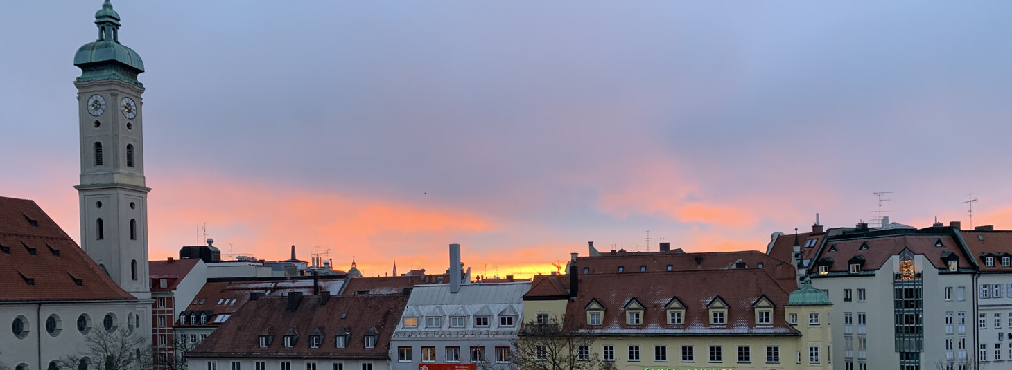 Blick auf Münchens Altstadt bei Sonnenuntergang, mit Kirchturm und historischen Gebäuden unter einem farbenfrohen Himmel.
