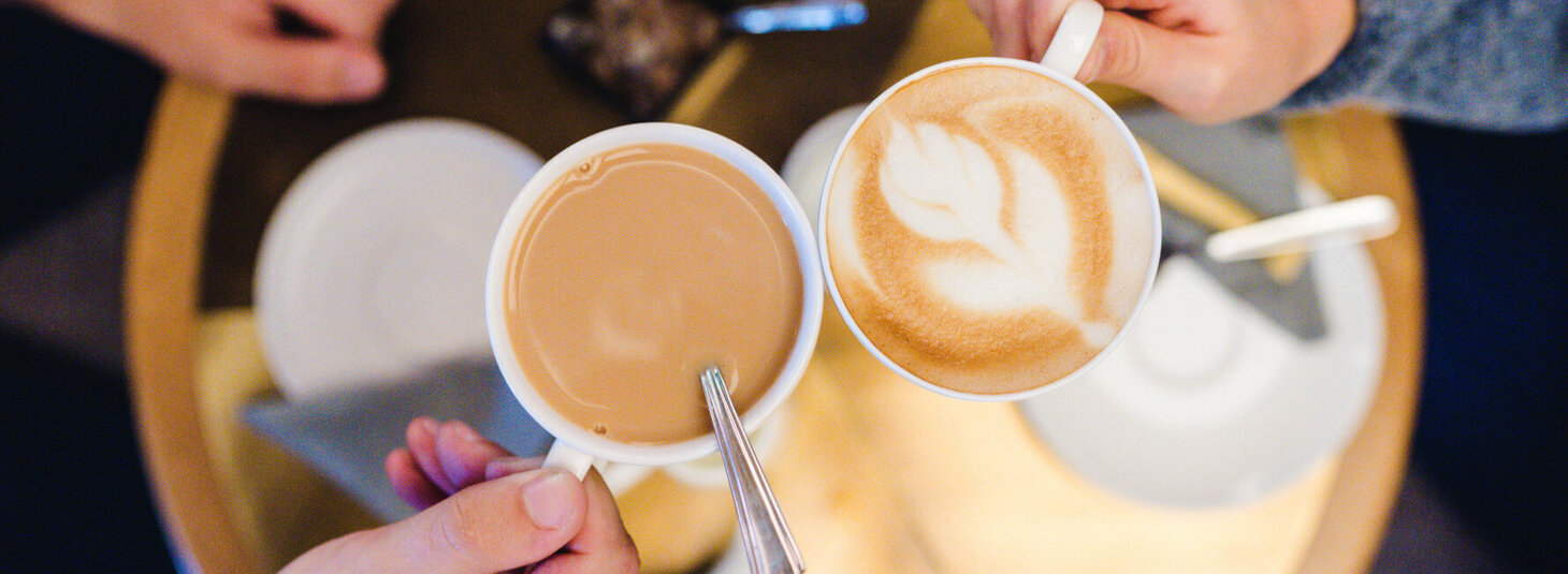 Zwei Kaffeetassen stoßen an, eine mit Latte Art, auf einem Holztisch im LOUIS Hotel München.
