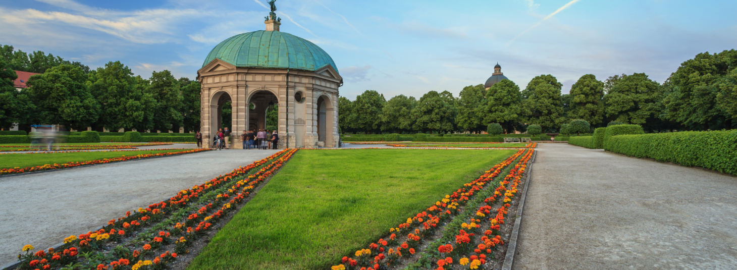 Pavillon im Hofgarten München, umgeben von Blumenbeeten und grünen Rasenflächen, unter blauem Himmel.