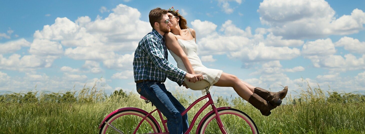 Ein Paar küsst sich auf einem pinken Fahrrad in einer grünen Wiese unter blauem Himmel mit weißen Wolken.