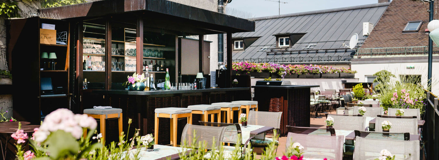 Dachterrasse des LOUIS Hotels mit Bar, Sitzplätzen und blühenden Pflanzen in München bei sonnigem Wetter.