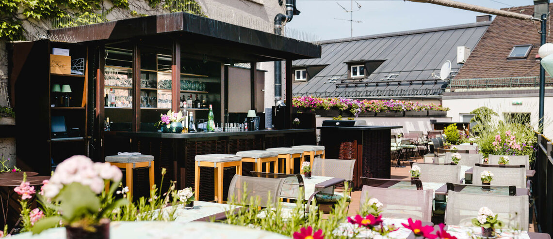 Dachterrasse des LOUIS Hotels mit Bar, Sitzplätzen und blühenden Pflanzen in München bei sonnigem Wetter.