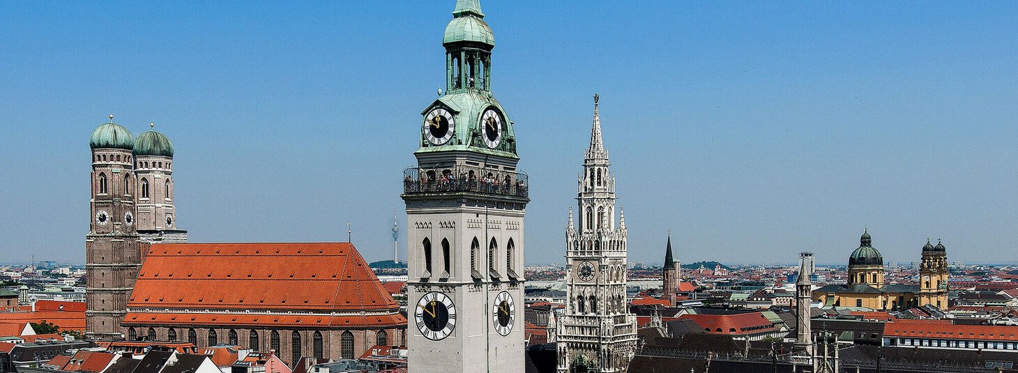 Stadtpanorama von München mit markanten Kirchtürmen und roten Dächern unter klarem, blauem Himmel.