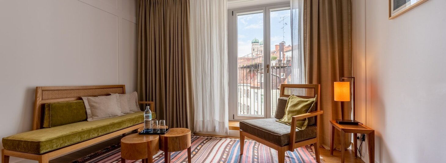 Gemütliches Hotelzimmer mit Retro-Möbeln, Teppich, Lampe und Balkonblick auf Münchens Architektur.