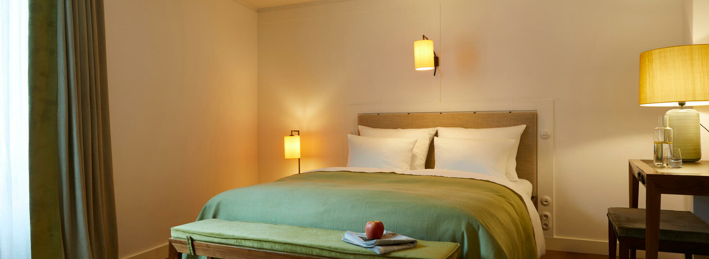 Gemütliches Hotelzimmer mit Doppelbett, grüner Tagesdecke, Holzbank, Schreibtisch und warmem Licht.