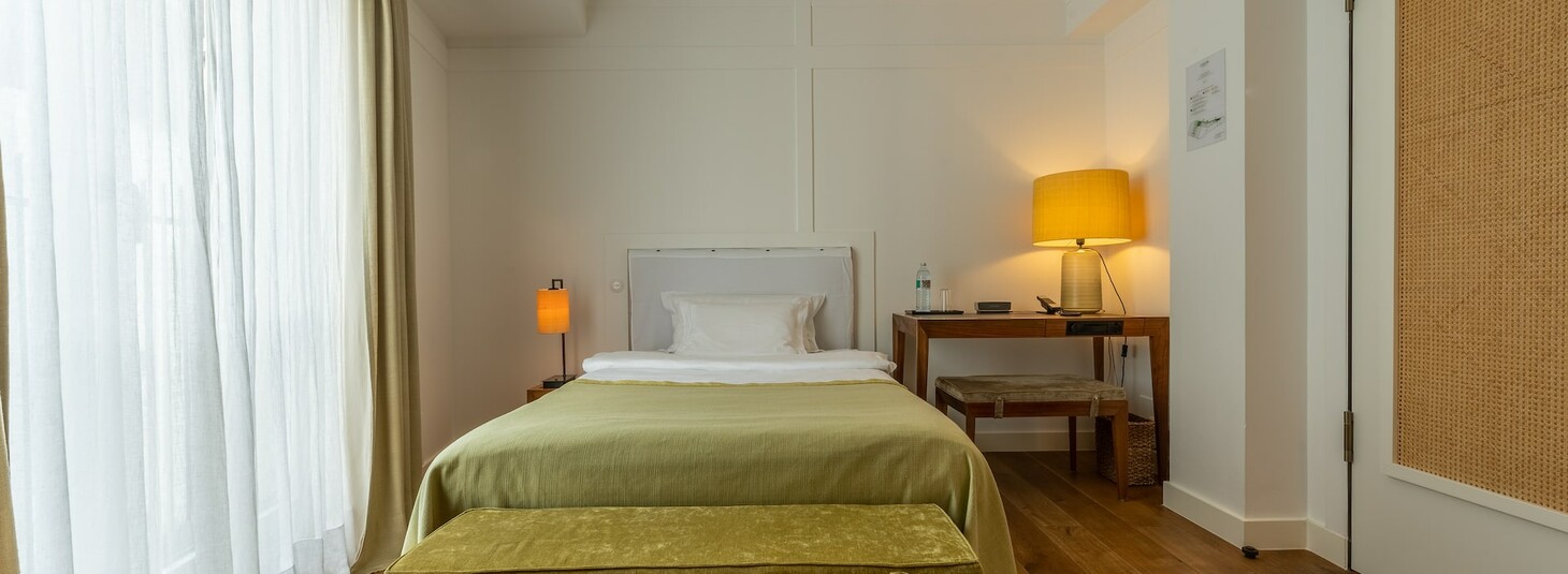 Helles Hotelzimmer mit Einzelbett, grünem Bettüberwurf, Schreibtisch, Lampen und großen Fenstern mit Vorhängen.