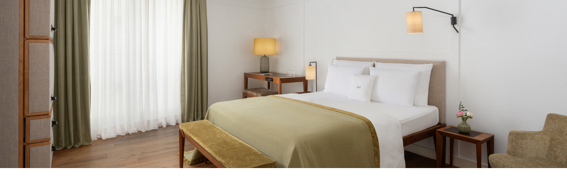 Cozy hotel room with double bed, green bedspread, wooden bench, desk and warm light.