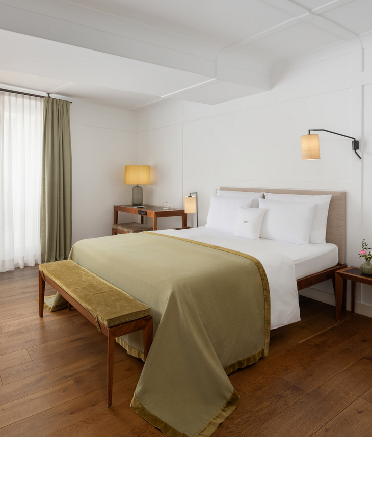 Cozy hotel room with double bed, green bedspread, wooden bench, desk and warm light.
