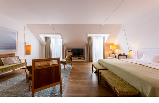 Bright hotel room with wooden floor, double bed, seating area, TV and large windows with curtains at the LOUIS Hotel Munich.