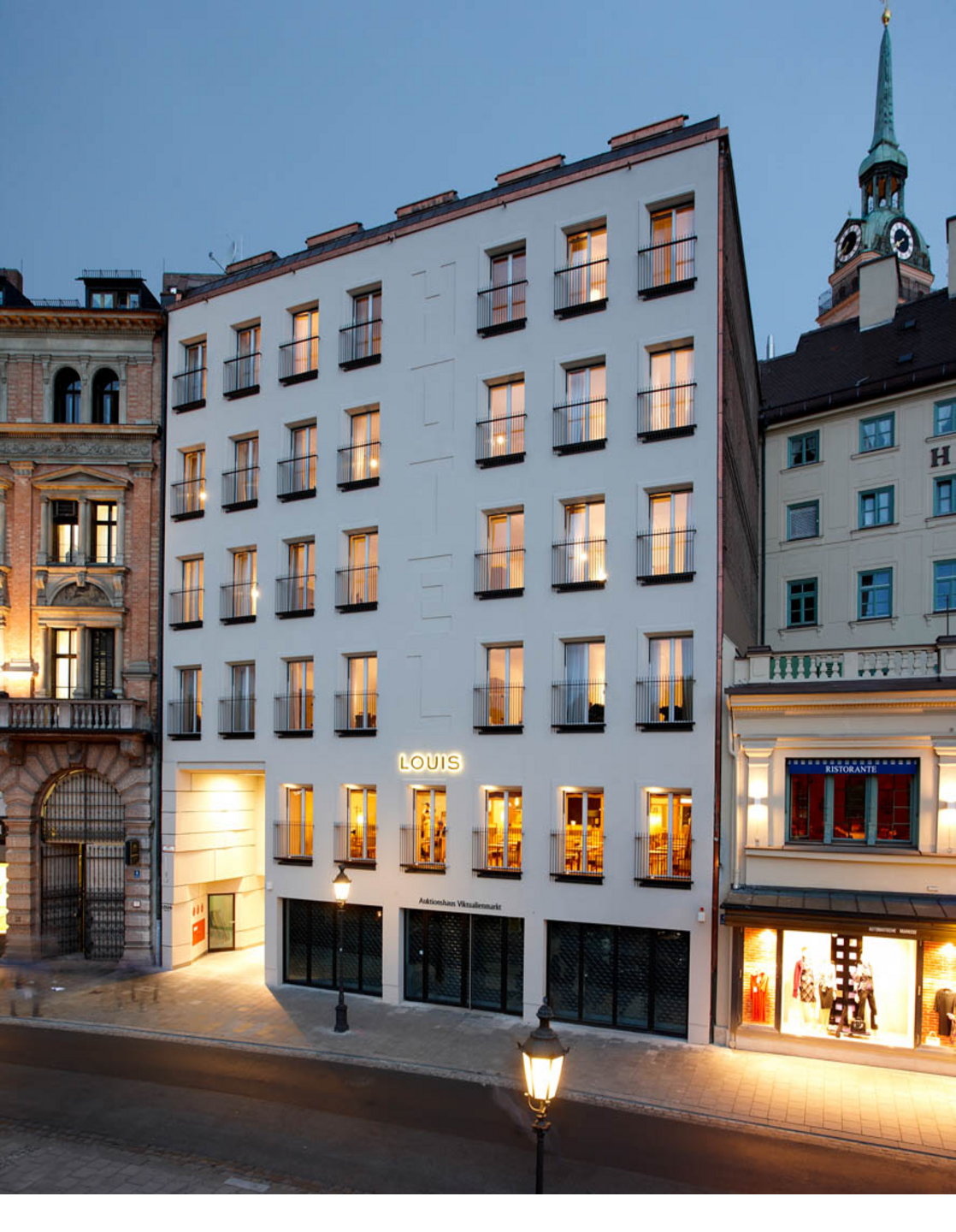 Exterior view of the LOUIS Hotel in Munich at dusk, with illuminated windows and surrounding buildings.