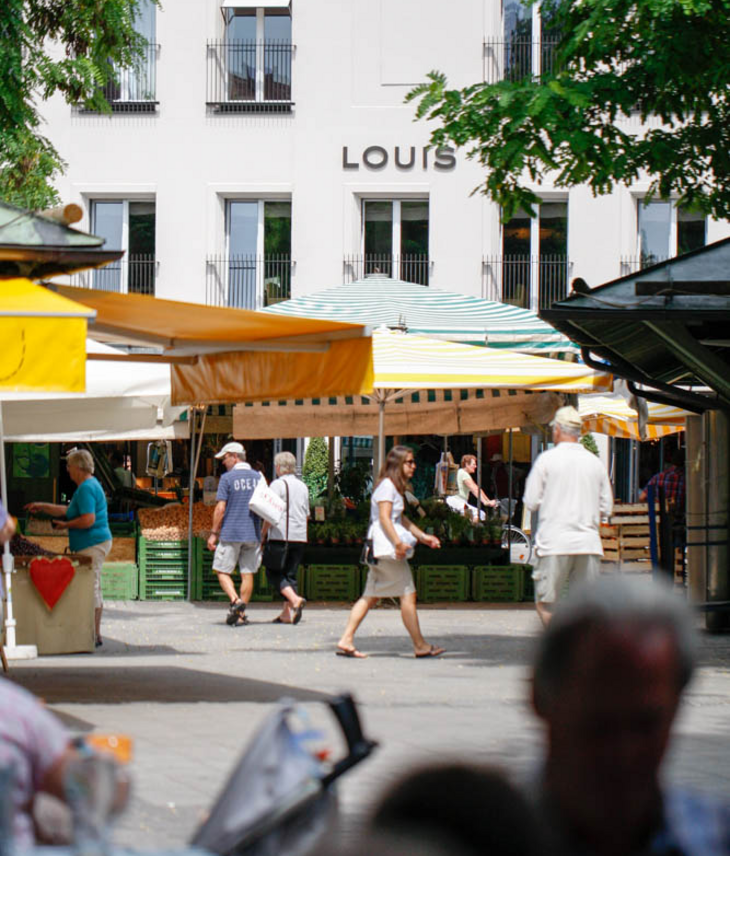 Blick auf das LOUIS Hotel München mit buntem Marktstand im Vordergrund und Passanten.