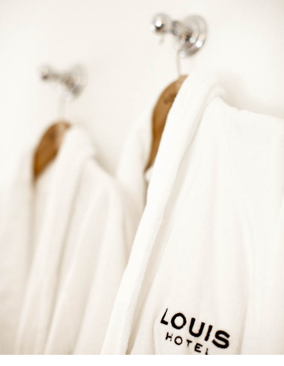 Two white bathrobes with the LOUIS Hotel logo hang on hooks on the wall.