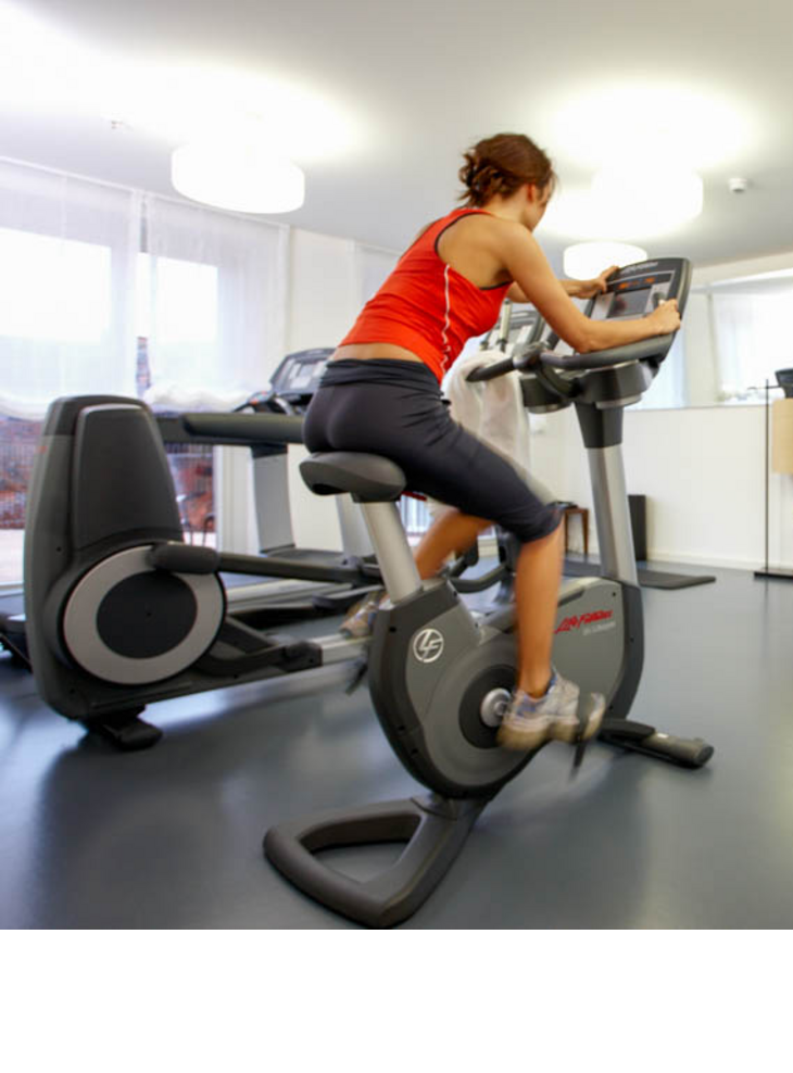 Woman training on an exercise bike in the fitness room of the LOUIS Hotel Munich, surrounded by fitness equipment.
