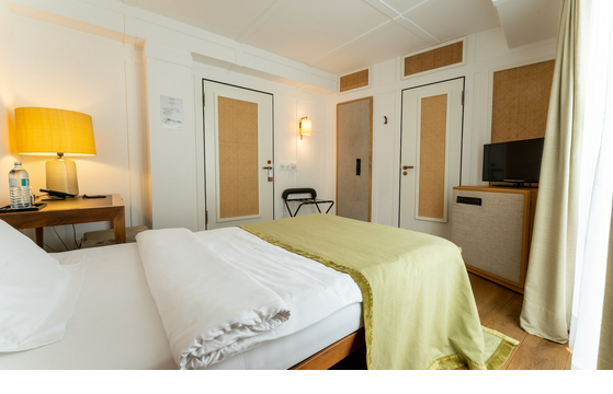 Helles Hotelzimmer mit gemütlichem Bett, Holzdetails, Schreibtisch, Lampe und kleinem Fernseher.