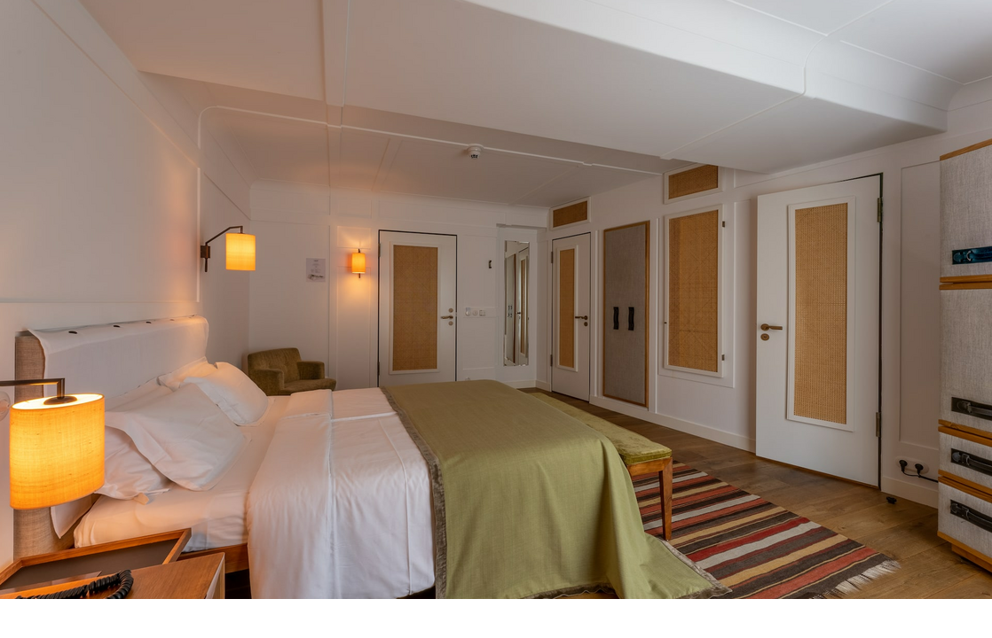 Helles Hotelzimmer mit Doppelbett, grüner Tagesdecke, Nachttischlampen, Teppich und Sessel im LOUIS Hotel München.