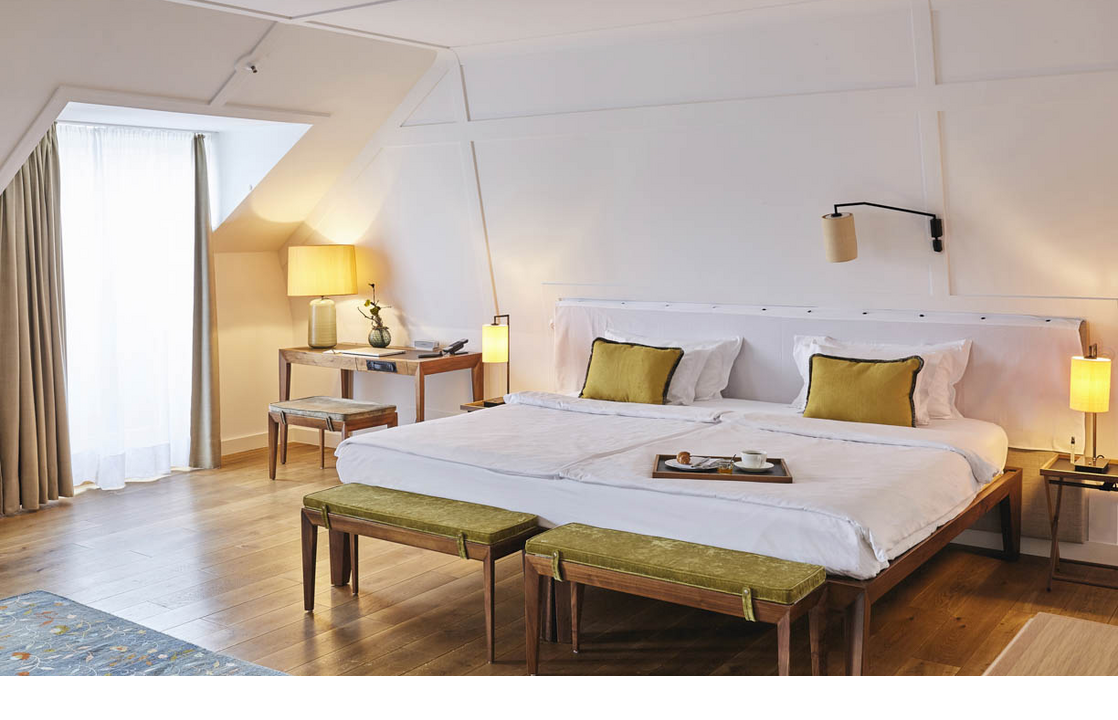 Helles Hotelzimmer mit großem Bett, Holzboden, Schreibtisch, zwei Hockern und einem Fenster mit Vorhängen.