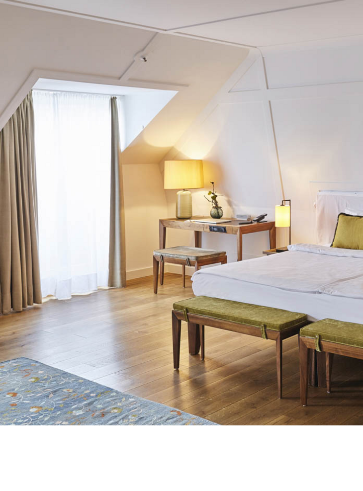 Bright hotel room with large bed, wooden floor, desk, two stools and a window with curtains.