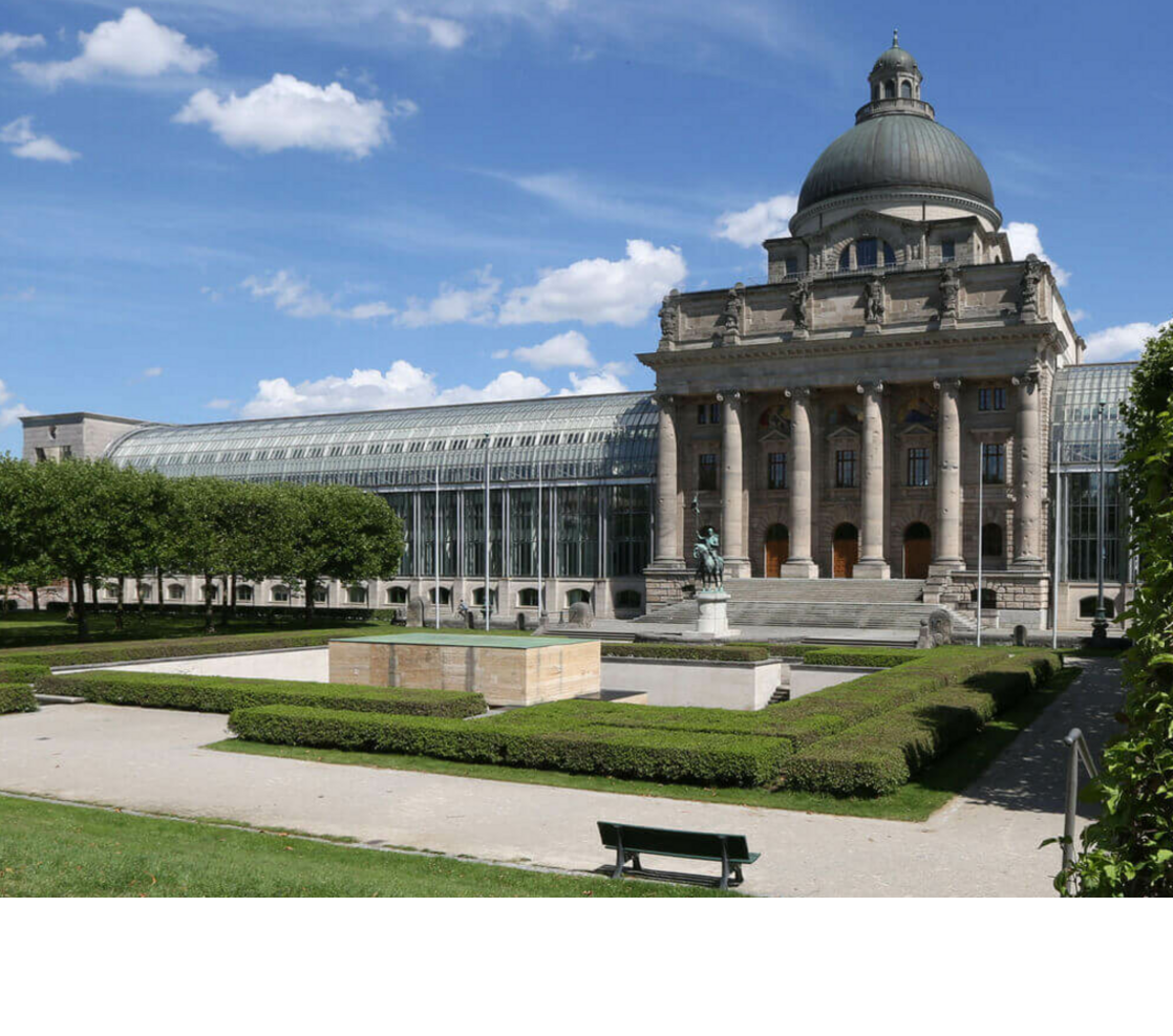 Historisches Gebäude mit Kuppel und Säulen, umgeben von gepflegten Gärten und Hecken unter blauem Himmel.