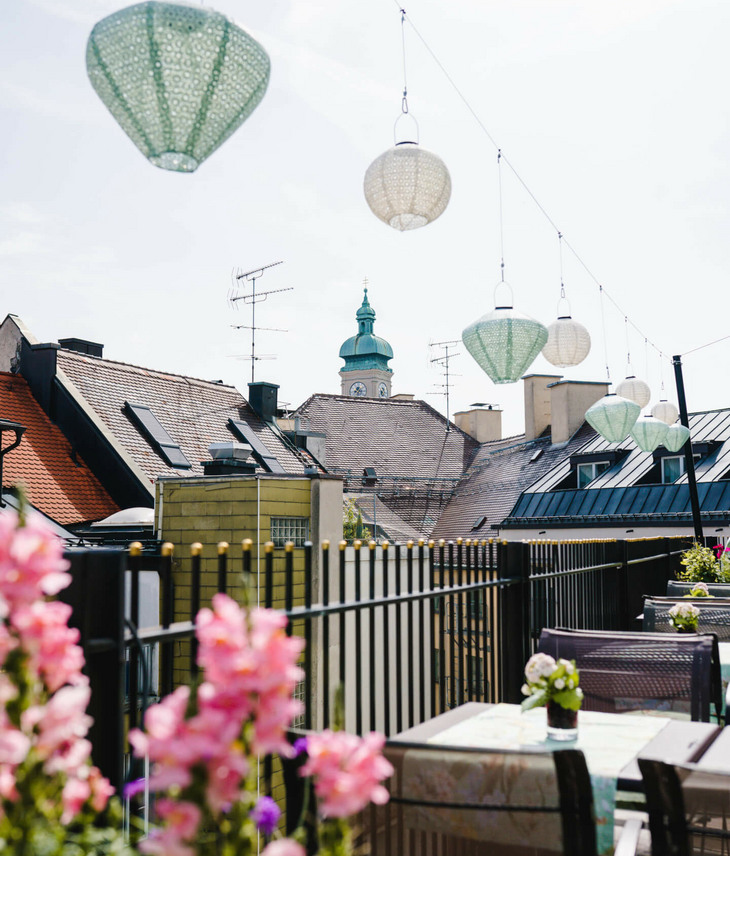 Blick von der Hotelterrasse mit Blumen, Hängelampen und Kirchturm im Hintergrund.