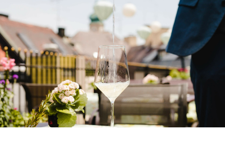 White wine is poured into a glass on a sunny hotel terrace, with flowers and city views in the background.
