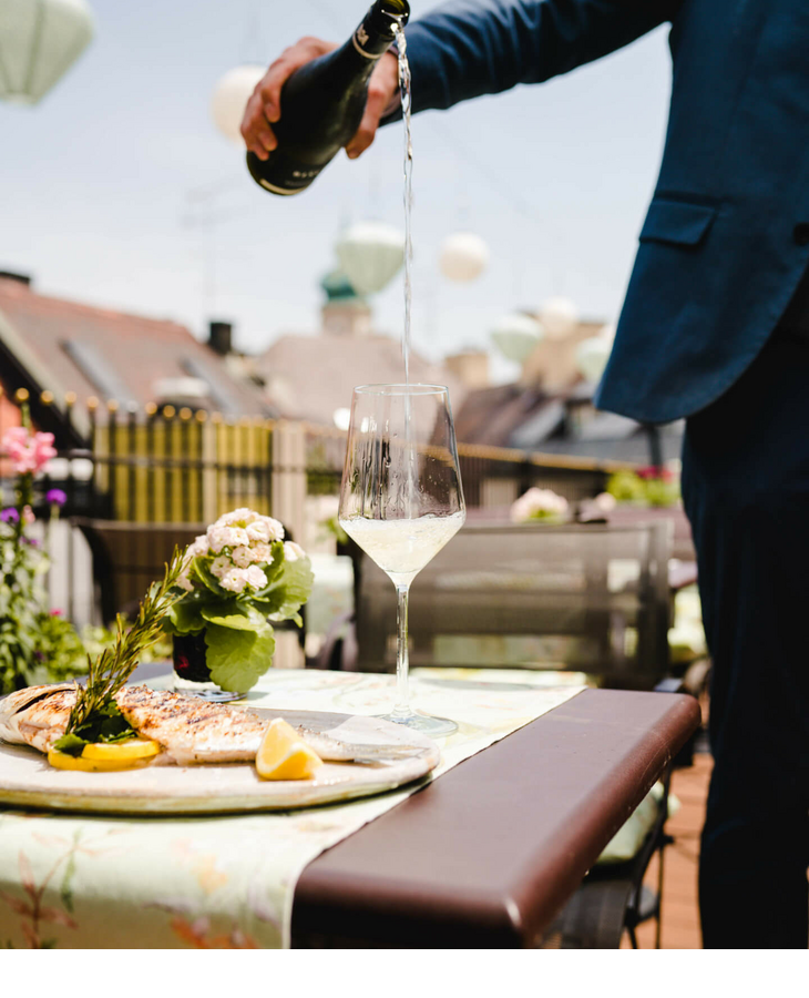 The Roof Terrace | Kühle Drinks auf der Dachterrasse des LOUIS Hotel München Person in Anzug schenkt Sekt auf sonniger Hotelterrasse ein, daneben ein Teller mit gegrilltem Fisch und Zitrone.