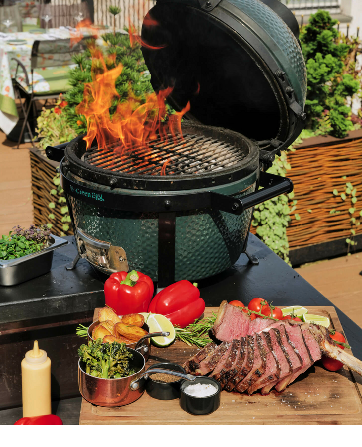 Barbecue with open flames, in front of it a wooden board with grilled meat, peppers and side dishes on a terrace.