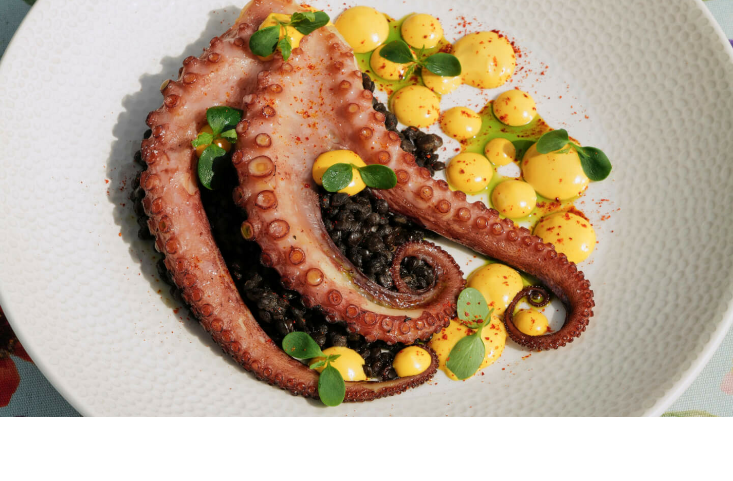 Grilled octopus on black rice with yellow sauce, served on a flowery table.