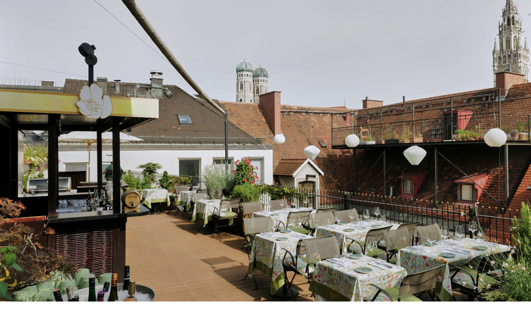 The Roof Terrace – Dachterrasse mit herrlichem Blick | LOUIS Hotel München Dachterrasse des LOUIS Hotels München mit gedeckten Tischen, Pflanzen und Blick auf die Türme der Frauenkirche.