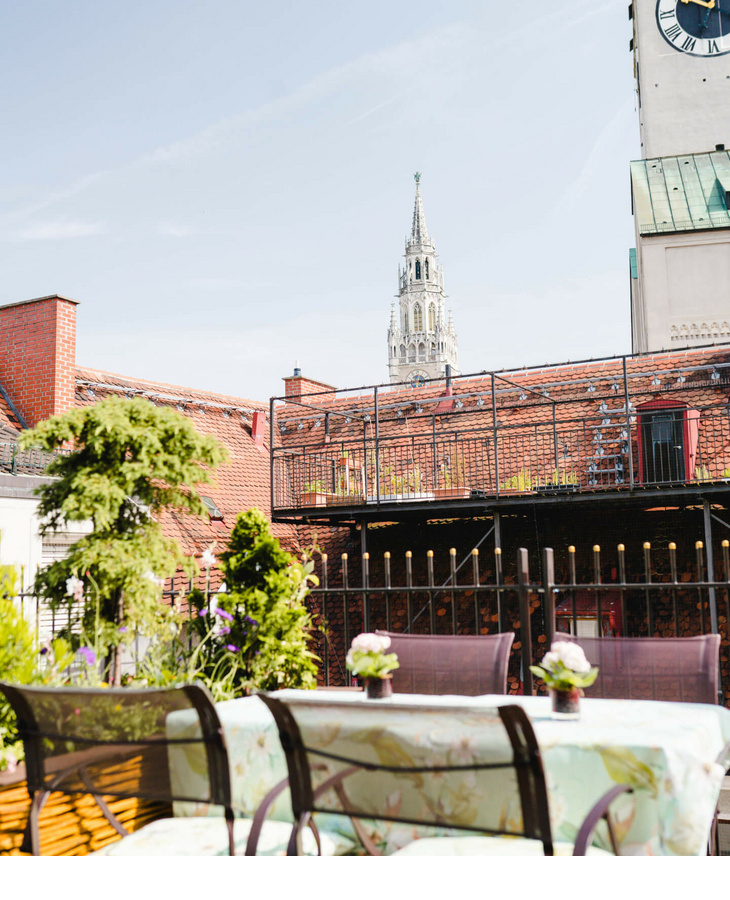 Dachterrasse mit Sitzbereich und Pflanzen, Blick auf Münchens Kirchturm bei blauem Himmel.
