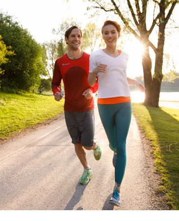 Zwei Personen joggen lachend auf einem sonnigen Parkweg, umgeben von Bäumen und Grünflächen.
