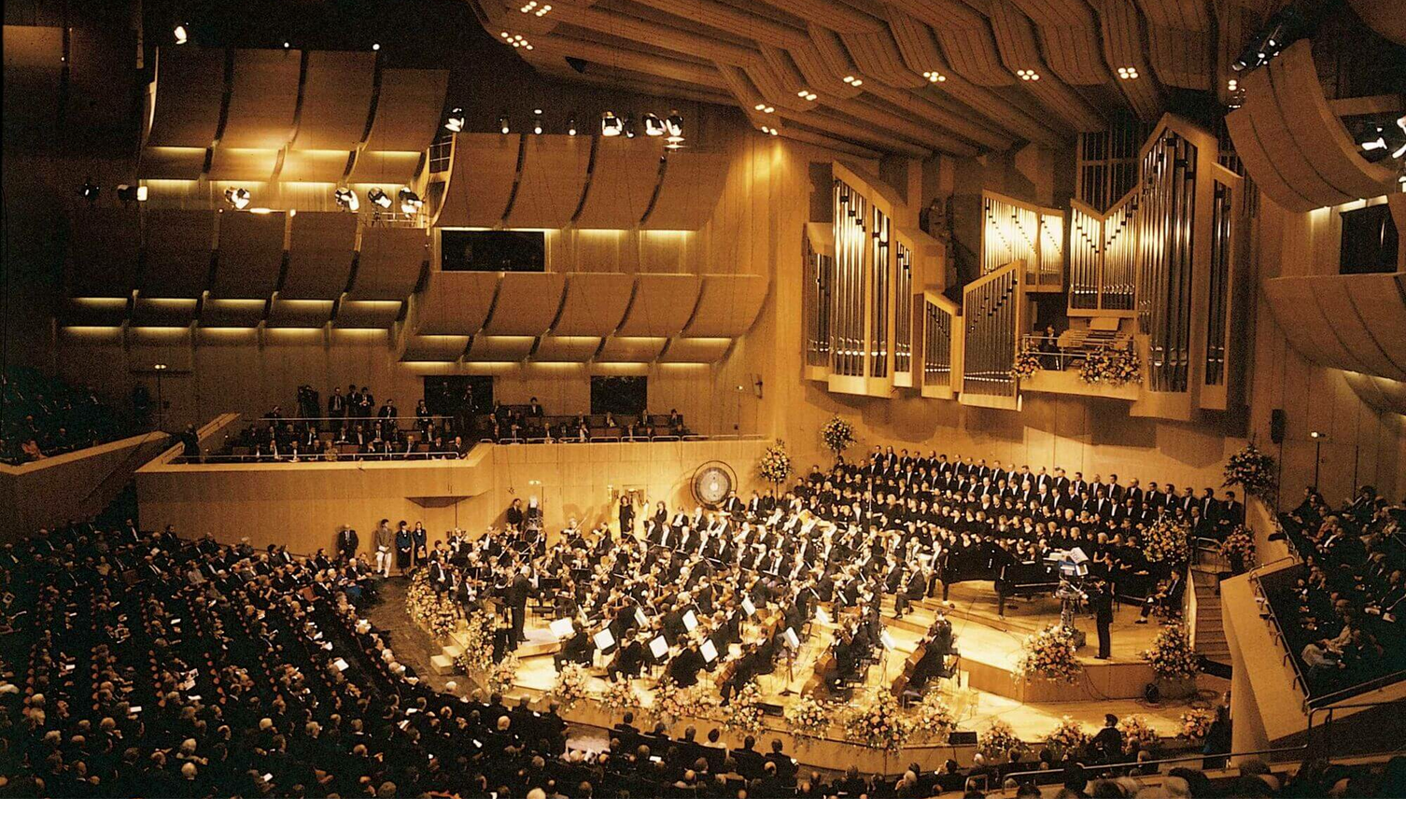 A concert hall with orchestra and choir on stage, surrounded by an audience in a modern, wood-paneled room.