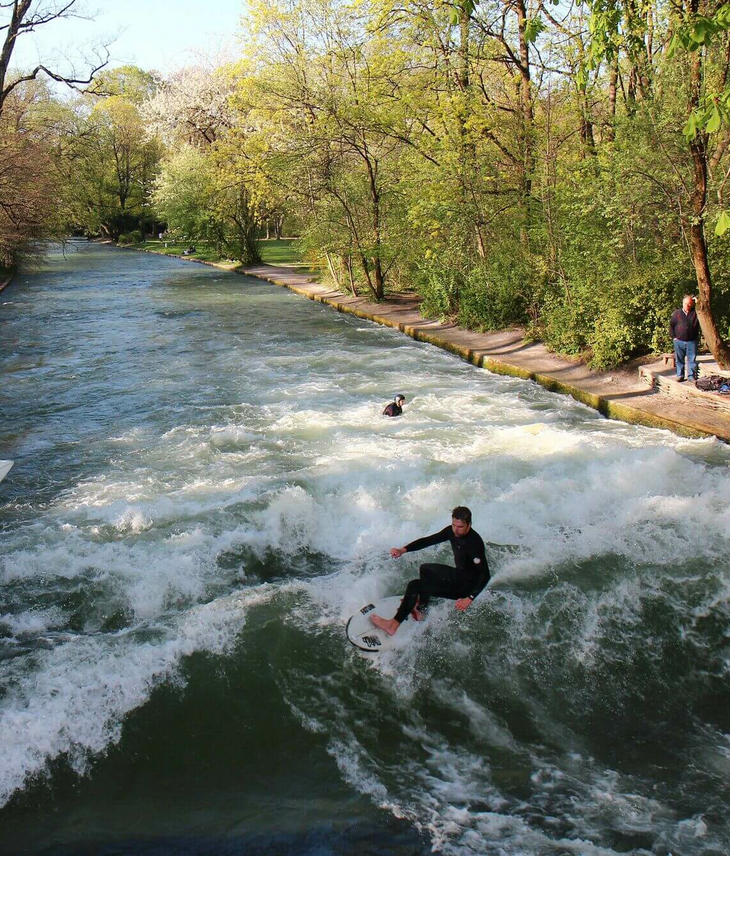 Surfer reiten auf einer stehenden Flusswelle, umgeben von Bäumen und Zuschauern am Ufer.