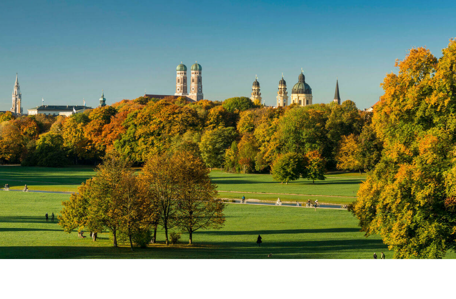 Weitläufiger Park mit grünen Wiesen, bunten Herbstbäumen und einer Stadtsilhouette im Hintergrund unter klarem Himmel.