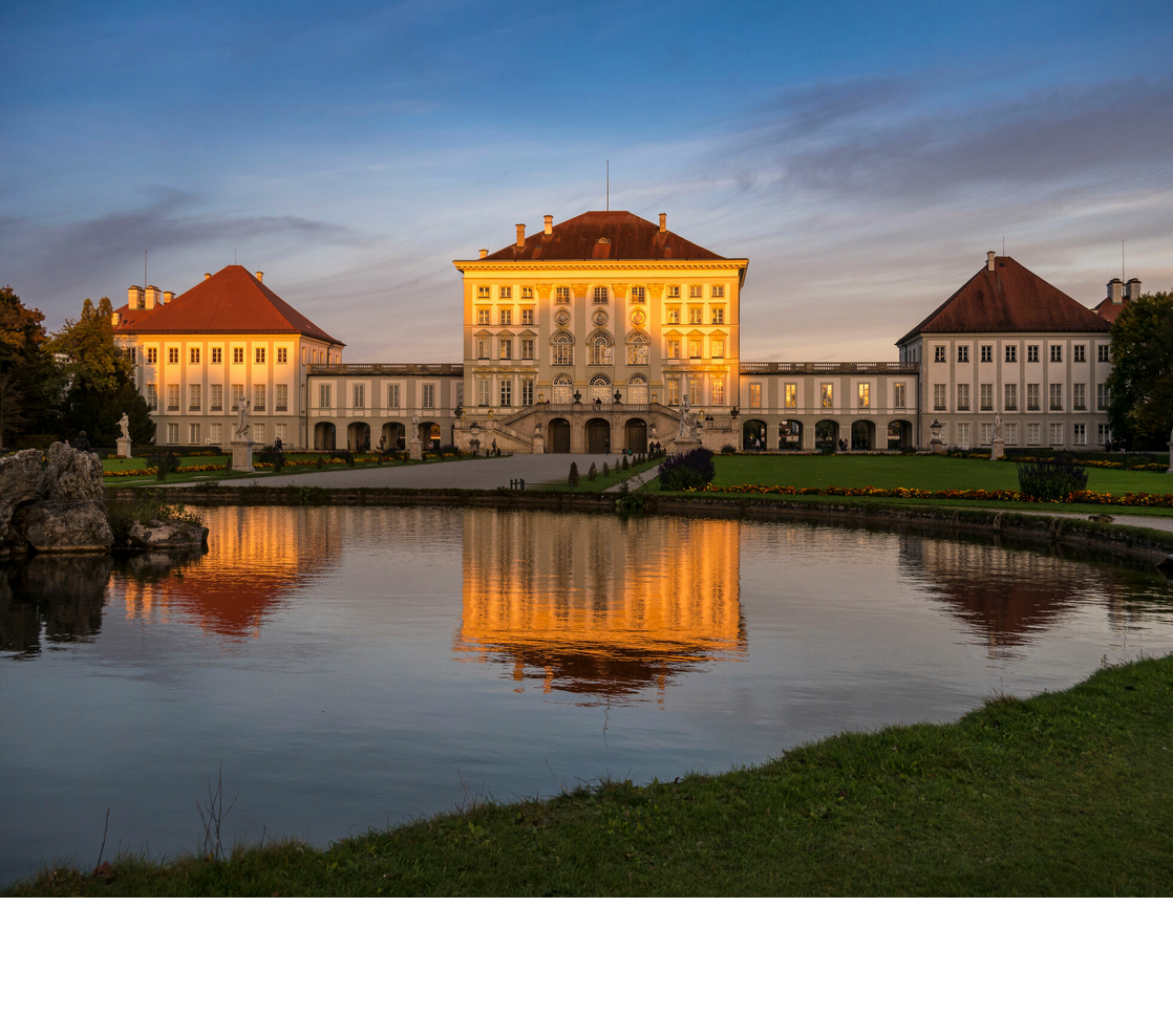 Historic castle with red roofs, surrounded by manicured gardens and a pond, reflected in the water at sunset.