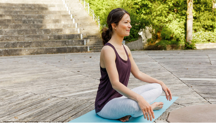 Frau macht Yoga im Freien auf einer blauen Matte, umgeben von Natur und Stufen im Hintergrund.