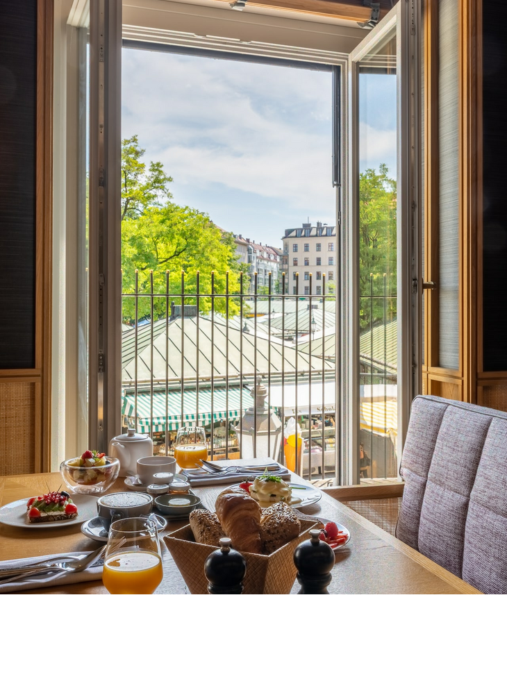 Breakfast table in the LOUIS Hotel with a view of the Viktualienmarkt through an open window.