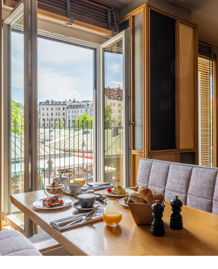 Frühstückstisch im LOUIS Hotel mit Blick auf Münchens Dächer durch ein geöffnetes Fenster.