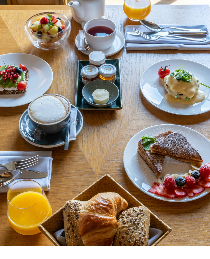 Frühstückstisch im LOUIS Hotel München mit Gebäck, Obst, Kaffee, Tee und Saft, elegant angerichtet auf Holztisch.