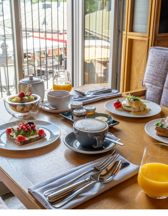 Frühstückstisch mit Kaffee, Saft, Obst und belegtem Brot im LOUIS Hotel München, mit Blick auf den Markt.