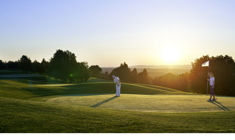Golfer auf grünem Platz bei Sonnenuntergang, eine Person hält die Fahne am Loch.