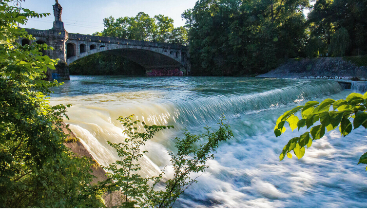 River with a small waterfall, a historic bridge above it, surrounded by lush greenery and trees.