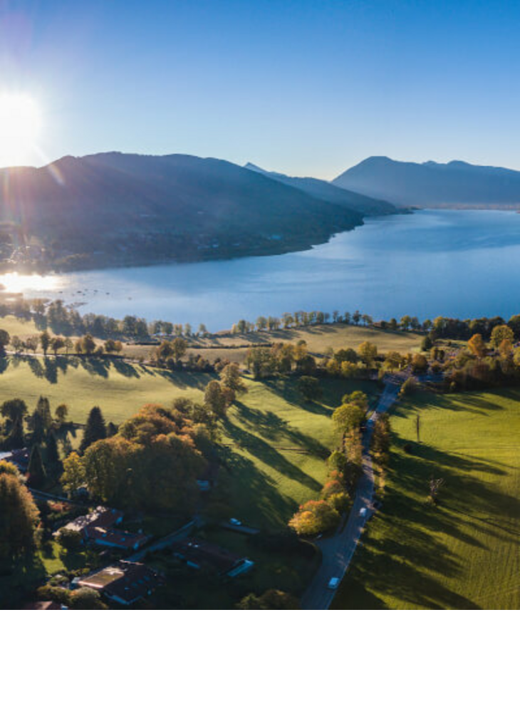 Severin*s Resort & Spa Tegernsee Aerial view of a lake with surrounding green meadows and mountains in the background at sunrise.